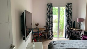 A hotel room with a bed, a TV mounted on the wall, a small table with a vase, a chair, a table with a lamp, and a sliding glass door leading to a balcony.
