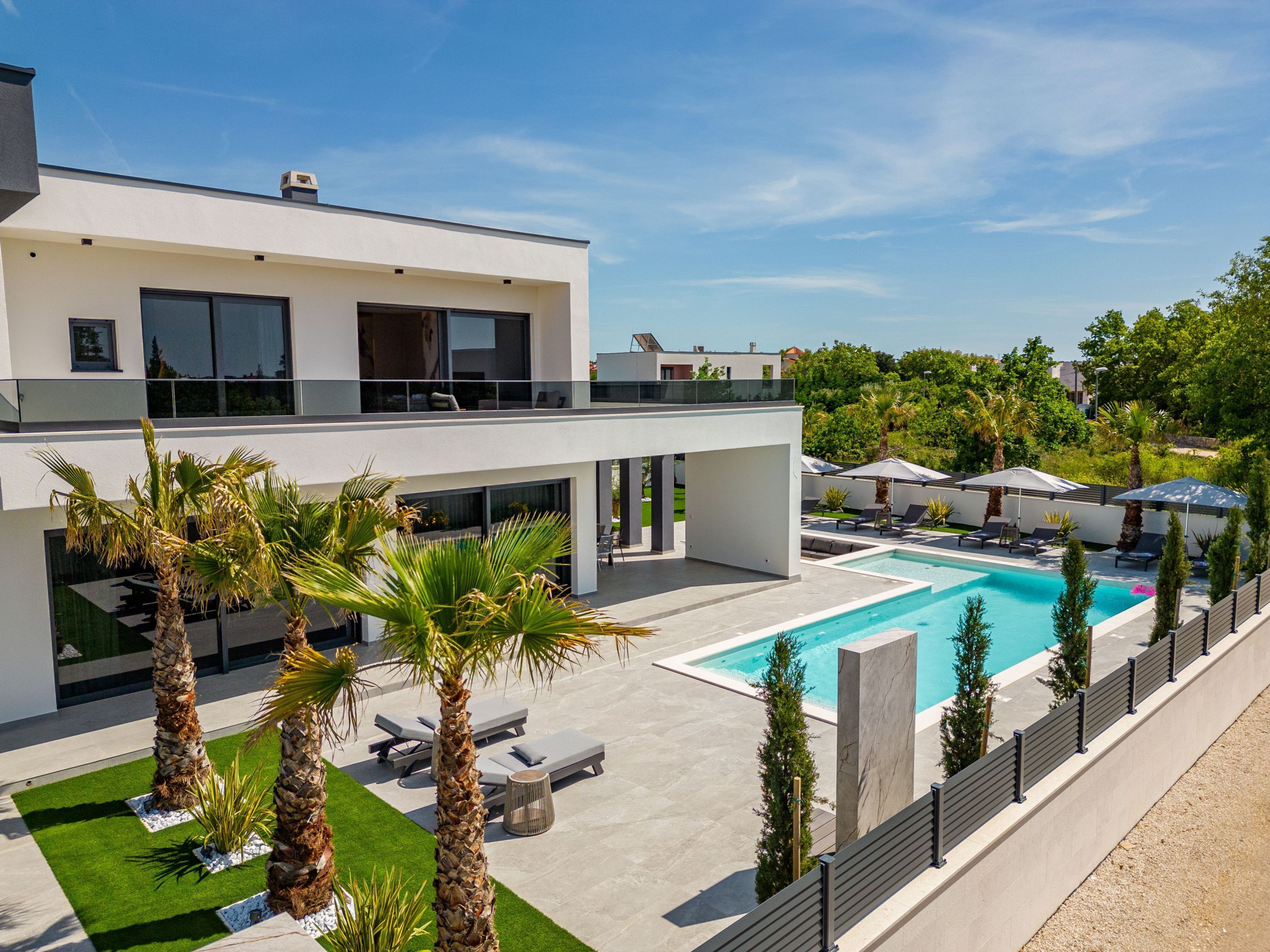 A modern two-story house with a swimming pool, outdoor seating, and palm trees.