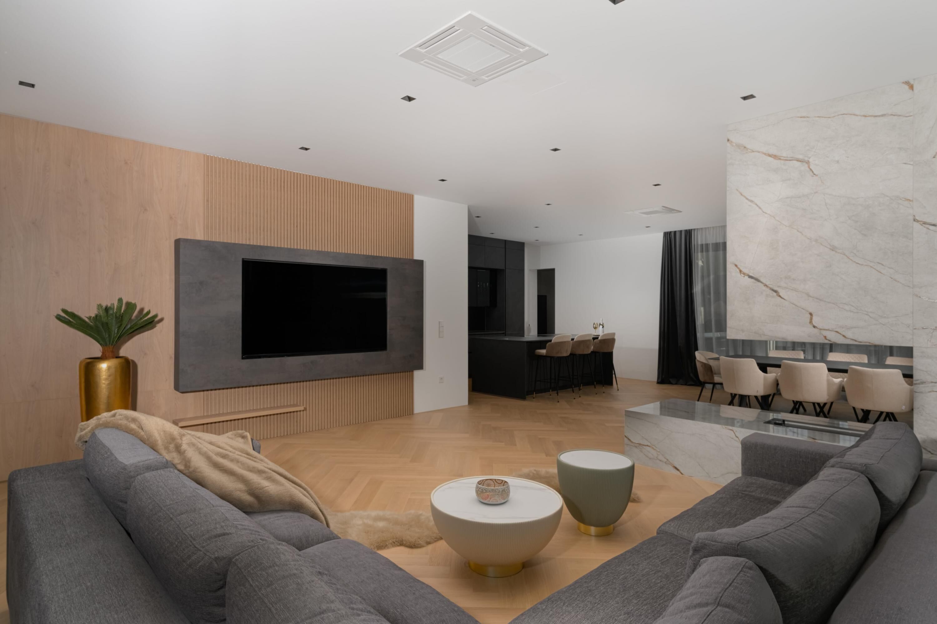 A modern living room with a large flat-screen TV, grey sofas, a coffee table, and a dining area with chairs.