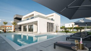 Une maison moderne à deux étages, dotée d’une piscine, d’un espace salon extérieur et d’un grand parasol.