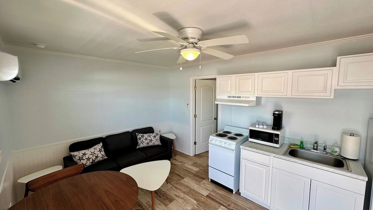 A compact kitchen with white cabinets, a stove, and a microwave. There's a ceiling fan, a small dining table, and a black sofa with patterned cushions.
