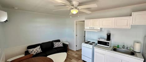 A compact kitchen with white cabinets, a stove, and a microwave. There's a ceiling fan, a small dining table, and a black sofa with patterned cushions.