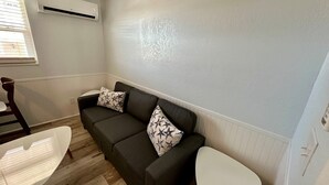 Una sala de estar pequeña y moderna, con un sofá gris oscuro, dos almohadones estampados, una mesa blanca y una ventana con persianas.