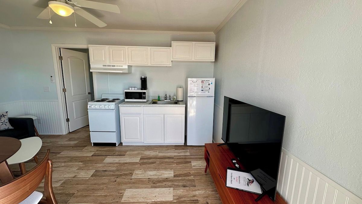 A compact kitchen with white cabinets, a stove, microwave, and refrigerator. There is a dining area with a table and chairs, and a flat-screen TV on a stand.