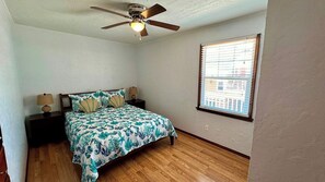 A bedroom with a bed, a ceiling fan, a window with blinds, and a nightstand with a lamp.