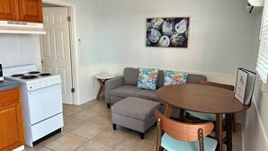 Een compacte keuken met een fornuis, een eettafel met stoelen, een grijze bank met kleurrijke kussens, en een schilderij van schelpen aan de muur.