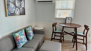 Een woonkamer met een grijze bank, kleurrijke kussens, een klein rond tafeltje en twee houten stoelen.