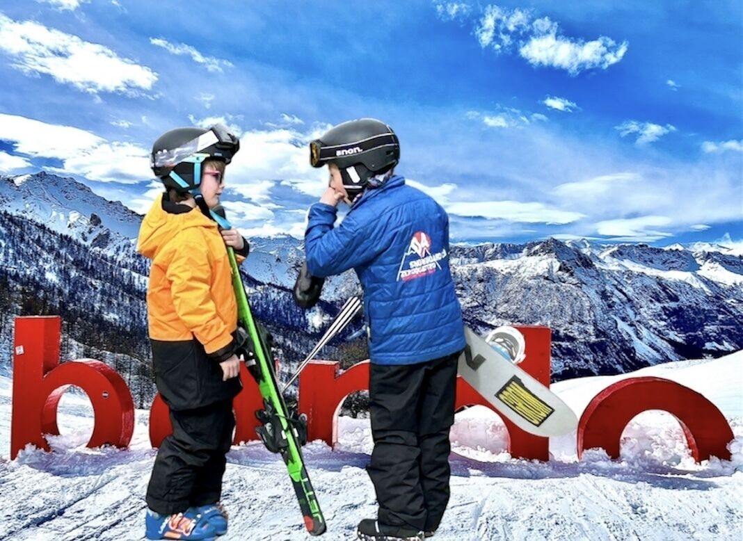 Twee personen op een besneeuwde berg met ski-uitrusting, staand voor grote rode letters die 'bode' spellen.