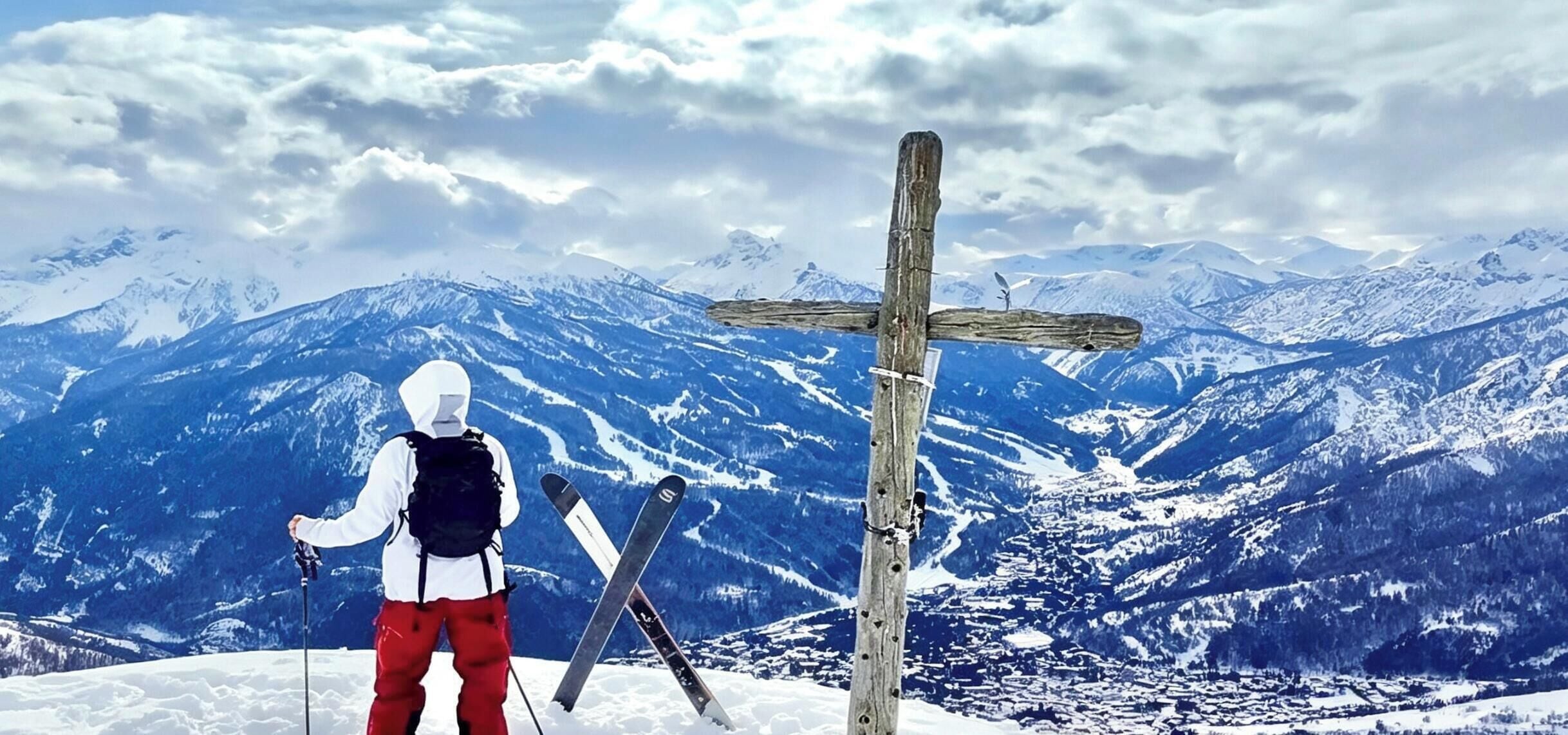 Een persoon met skiës en stokken staat in een besneeuwd landschap met bergen op de achtergrond.