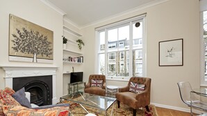 Una sala de estar con chimenea, dos sillones, una mesa de centro de vidrio y vistas a edificios a través de amplios ventanales.