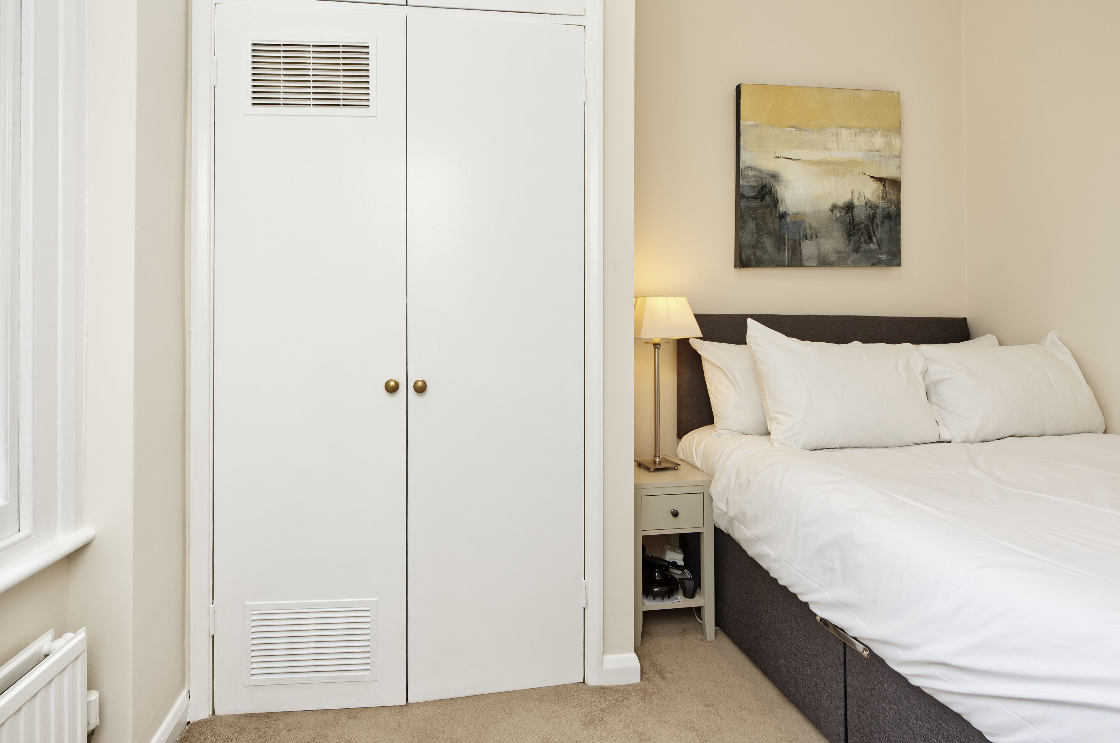 A bedroom with a bed, a nightstand, a lamp, a wardrobe, and a painting on the wall.