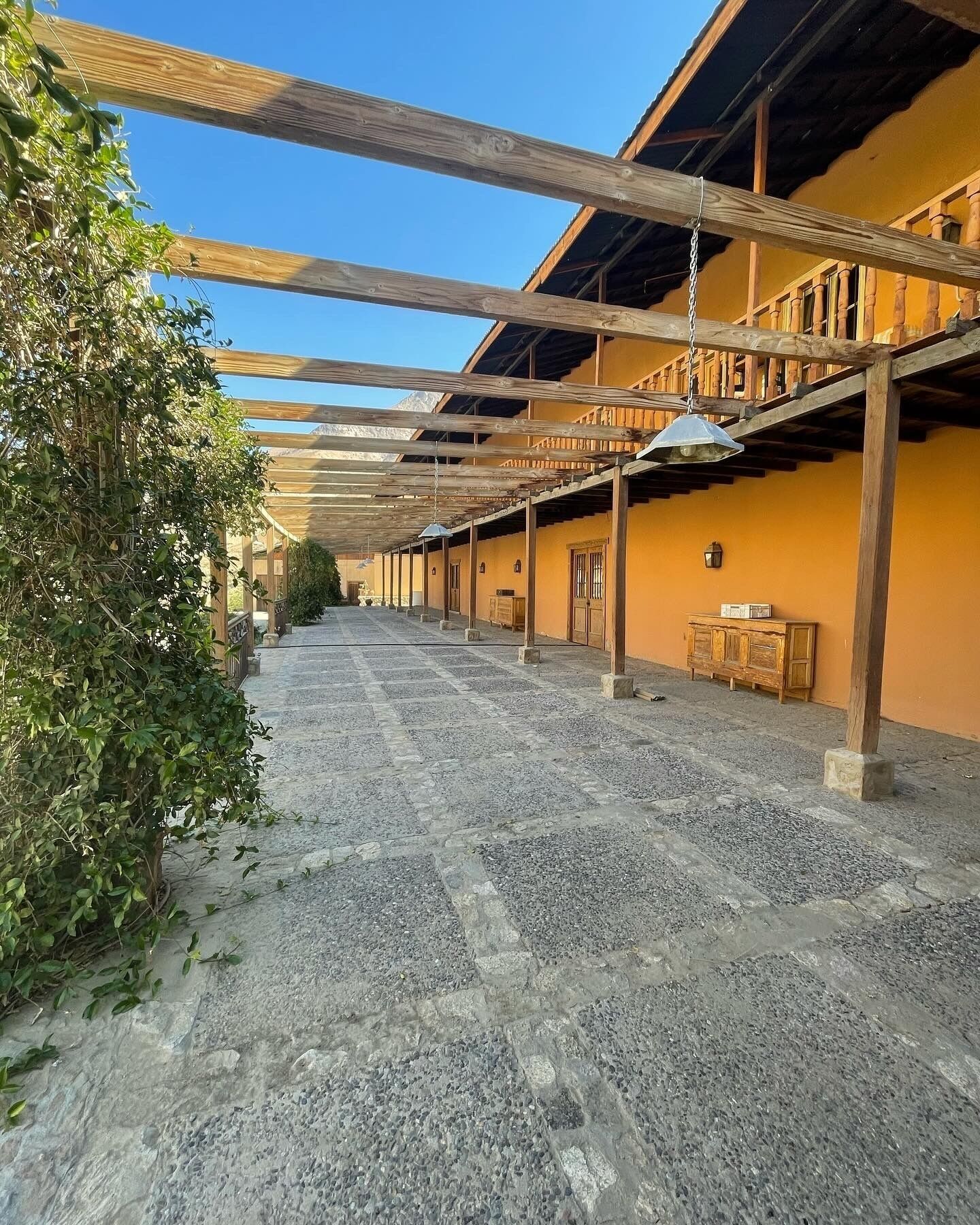 A covered outdoor area with a gravel floor, wooden beams, and a building with a yellow facade.