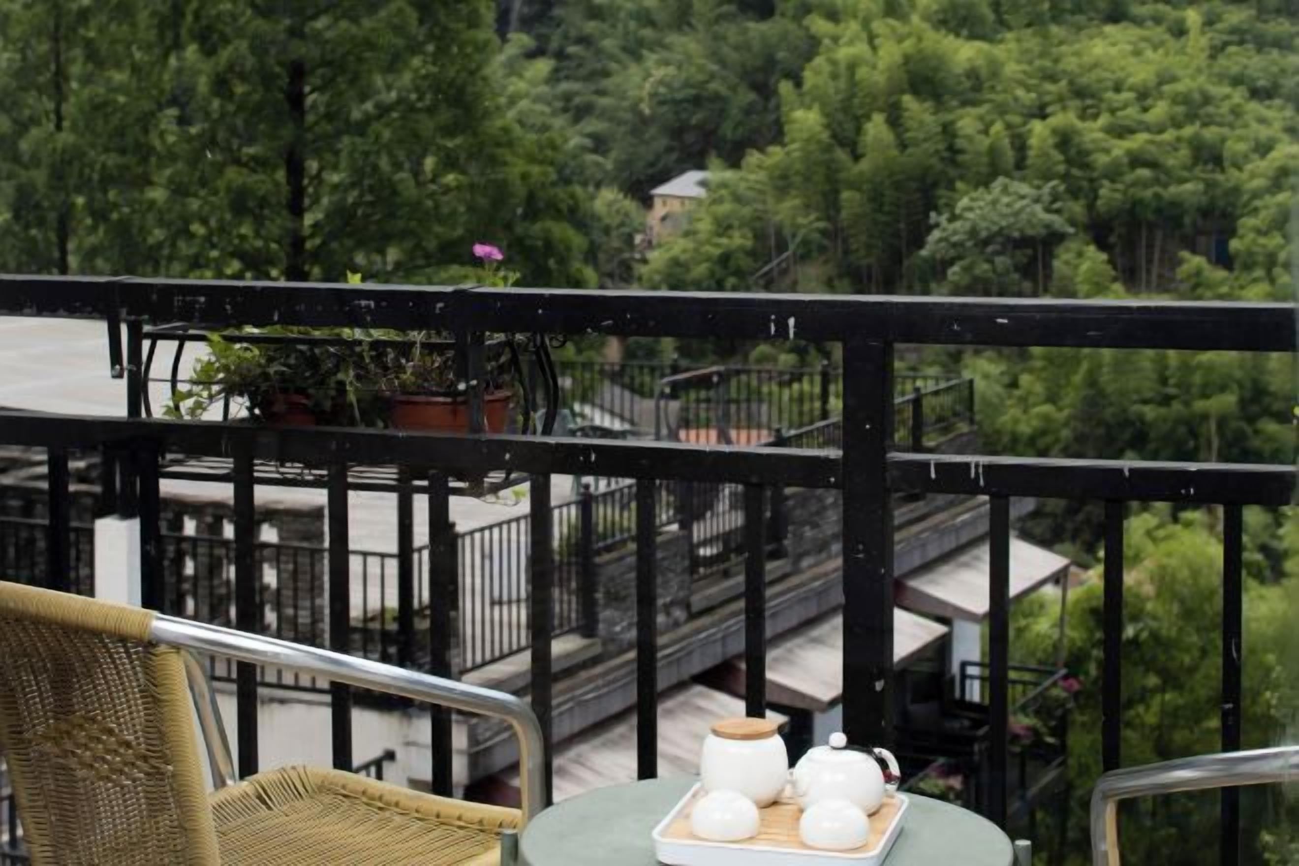 A balcony with a wicker chair, a small table with teapots and cups, and a view of a lush green forest.