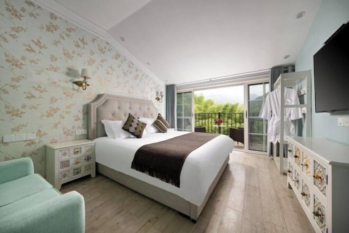 Een slaapkamer met een groot bed, een nachtkastje, een dressoir, een televisie en een balkon met uitzicht op groen.