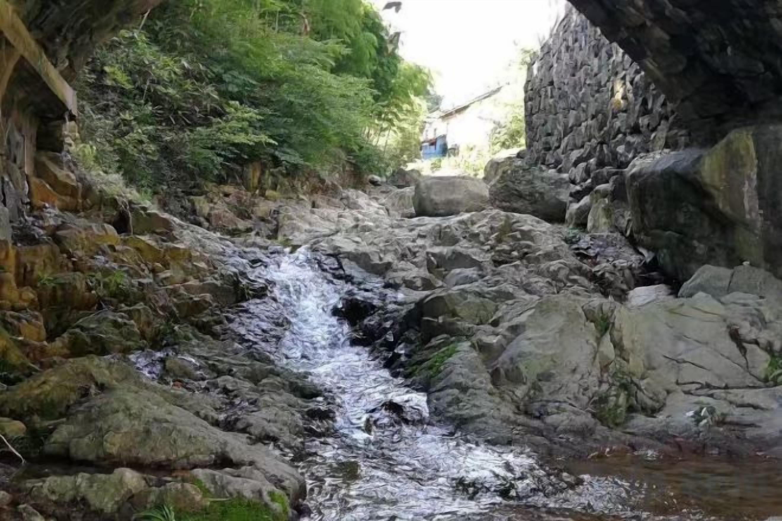 Ein schmaler Fluss fließt durch ein felsiges Gelände, an einer Seite eine Steinmauer und auf der anderen eine teilweise sichtbare Gebäude.