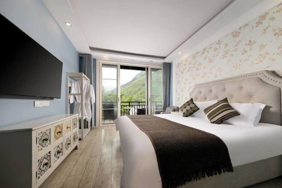 Een slaapkamer met een groot bed, een televisie, een kledingkast en een balkon met uitzicht op groen.