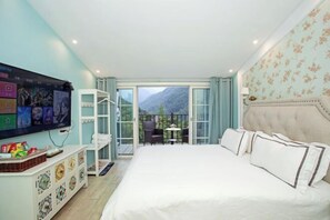 Une chambre avec un grand lit, un téléviseur fixé au mur, un balcon avec vue sur les montagnes et une petite table avec des chaises.