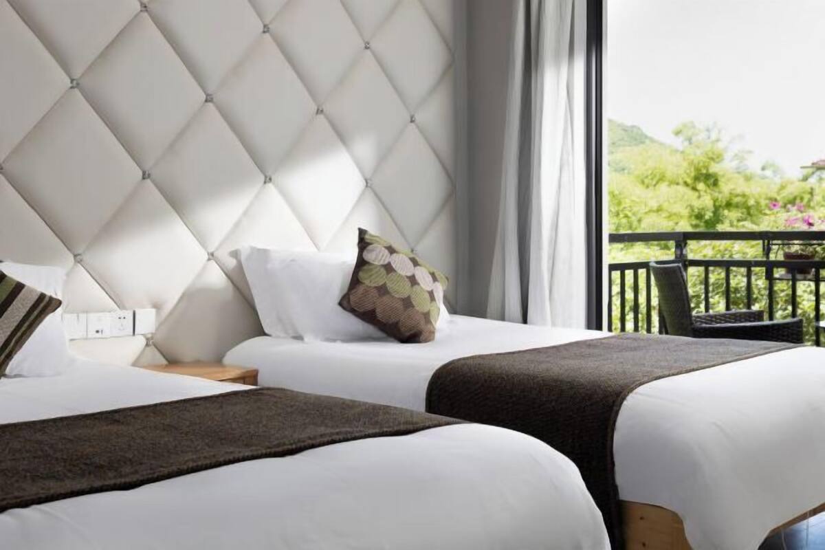 Een hotelkamer met twee bedden, een gestoffeerd hoofdeinde, een balkon met uitzicht op groen en een nachtkastje met een lamp.