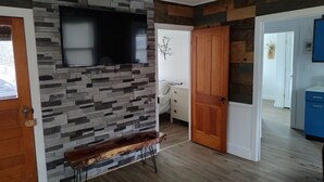 Una sala de estar moderna con una pared de piedra, un televisor montado, un banco de madera y una puerta que da a otra habitación.