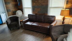 Un salón con un sofá de cuero marrón, una mesita auxiliar pequeña, una lámpara de pie y un sillón blanco mullido.