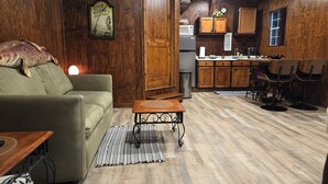 Una sala de estar acogedora con un sofá, una mesa de centro y un comedor con sillas. Se ve una cocina al fondo con gabinetes de madera y un microondas.