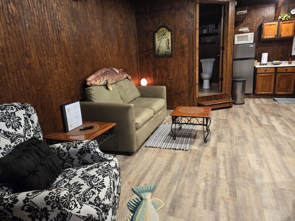 Un salon chaleureux avec un canapé, une table basse et un fauteuil à motifs. On aperçoit à l’arrière-plan un coin cuisine avec des meubles en bois et un four à micro-ondes.