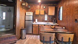 Cocina compacta con armarios de madera, un refrigerador de acero inoxidable, un microondas y un lavabo. Hay una zona de comedor con una mesa y sillas de madera.