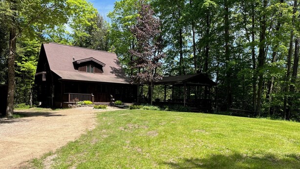 A rustic cabin with a large porch and a covered outdoor seating area, surrounded by a well-maintained lawn and dense trees.