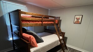 Una cama litera con escalera, una ventana con persianas, un cuadro en la pared y piso de madera.