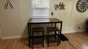 A room with a wooden table and two chairs, a window with blinds, a wall clock, and a wall decoration with the phrase "ONE TAKE TIME".