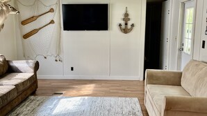 Una sala de estar con un sofá color beige, un tapete estampado, piso de madera, un televisor montado en la pared y una puerta que da a otra habitación.