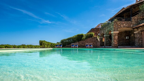 A clear pool with lounge chairs, a stone building with balconies, and a clear blue sky.