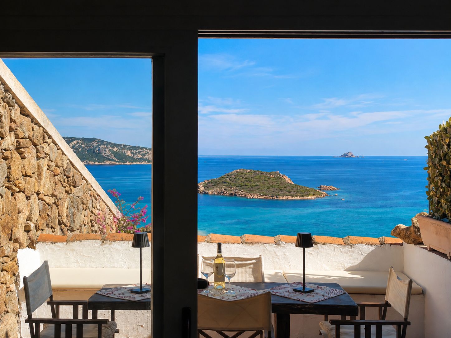 A terrace with a table and chairs, overlooking a coastal view with clear blue sea and distant islands.