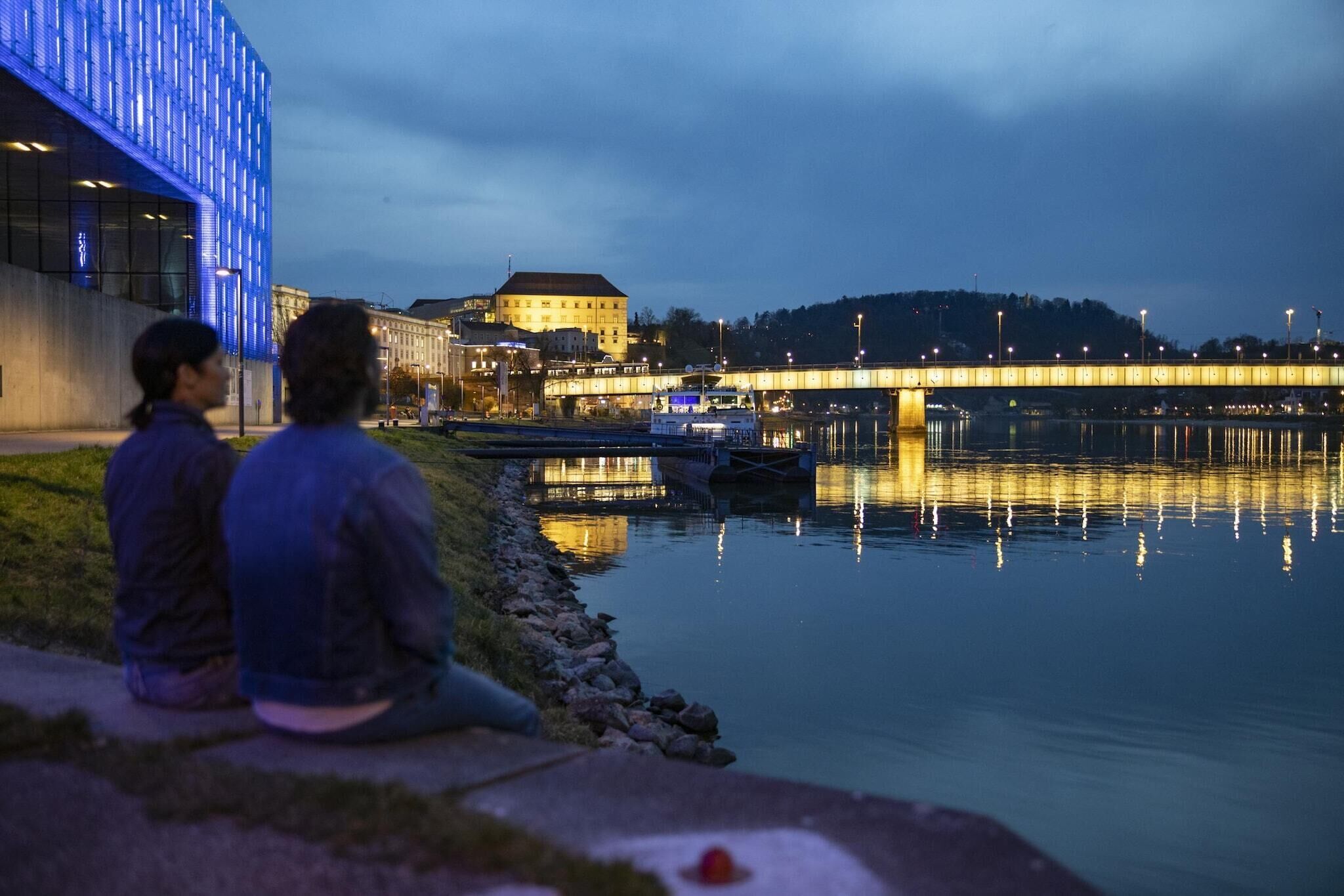 Two people sitting by a calm river with a bridge and buildings in the background.