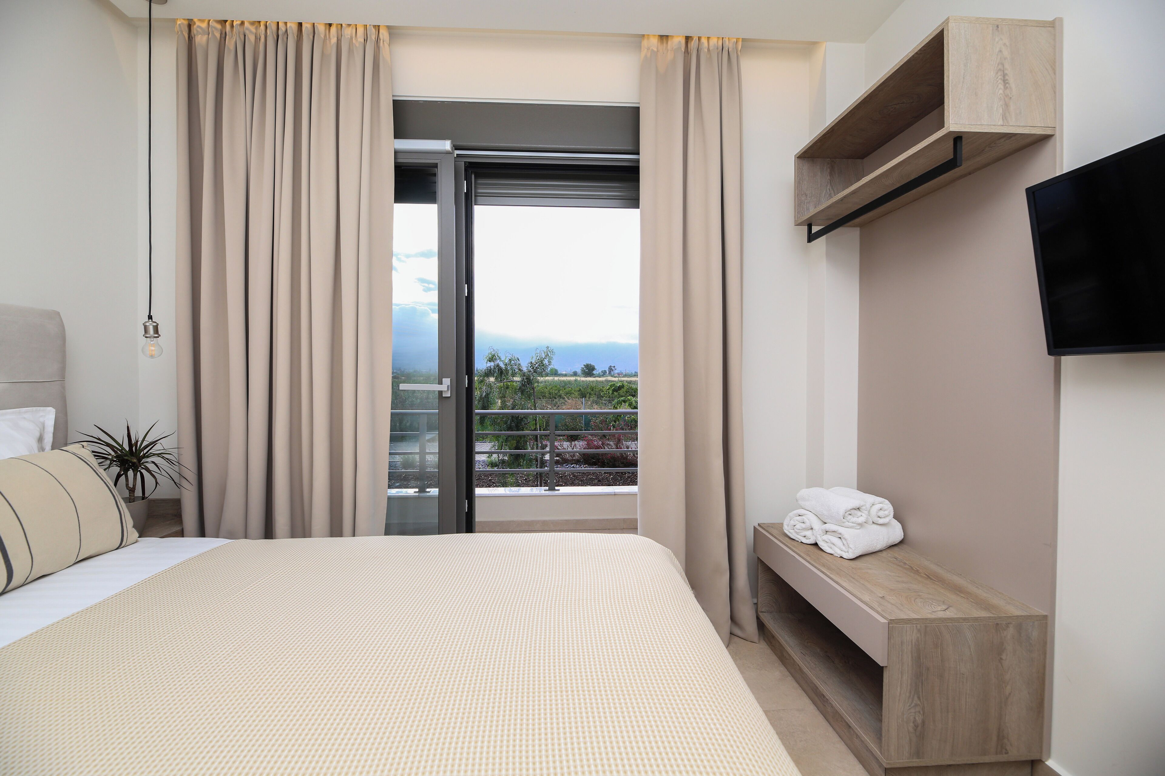 A hotel room with a bed, a TV mounted on the wall, a balcony with curtains, and a view of the outdoors.