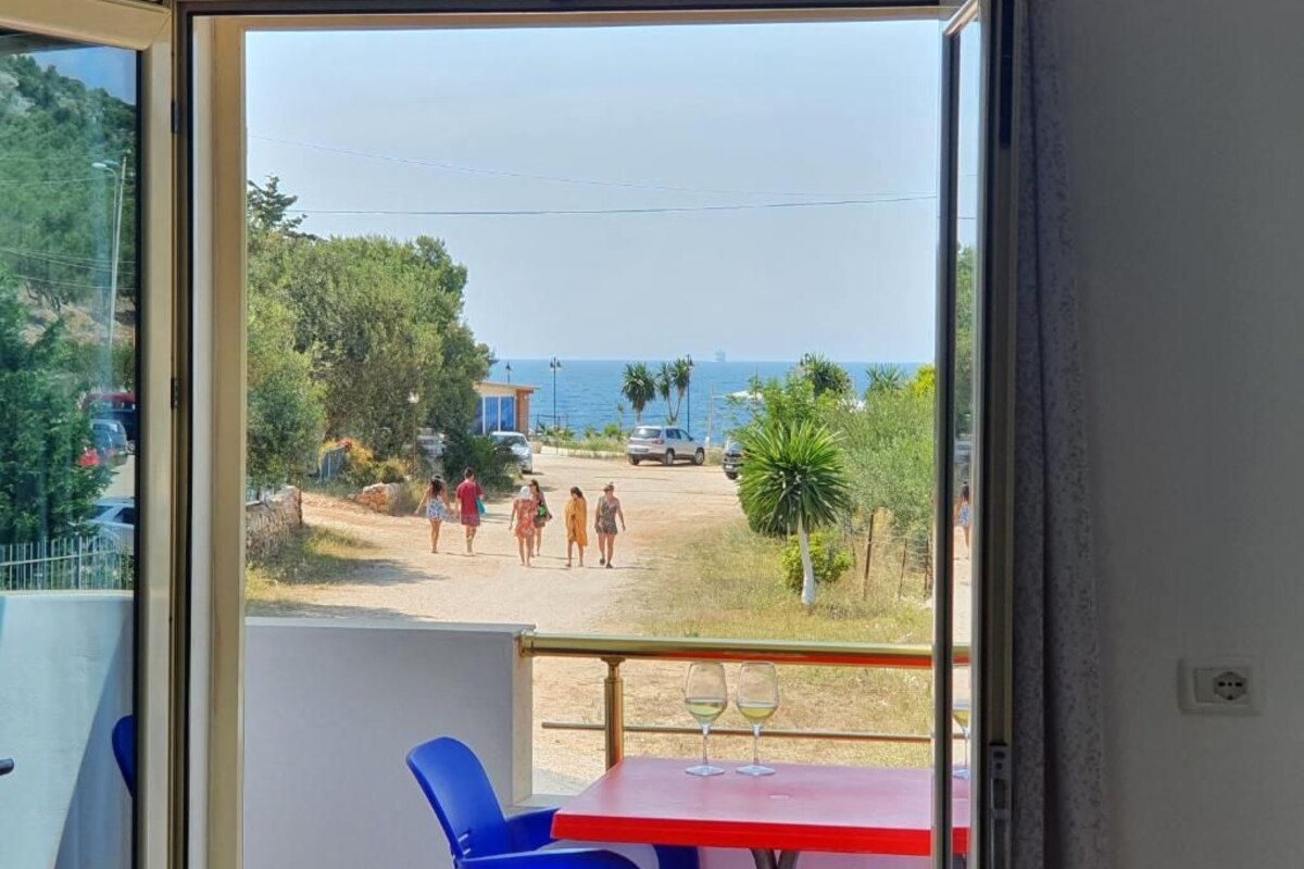 A balcony with a table and chairs, a view of the sea, and a road with people walking.