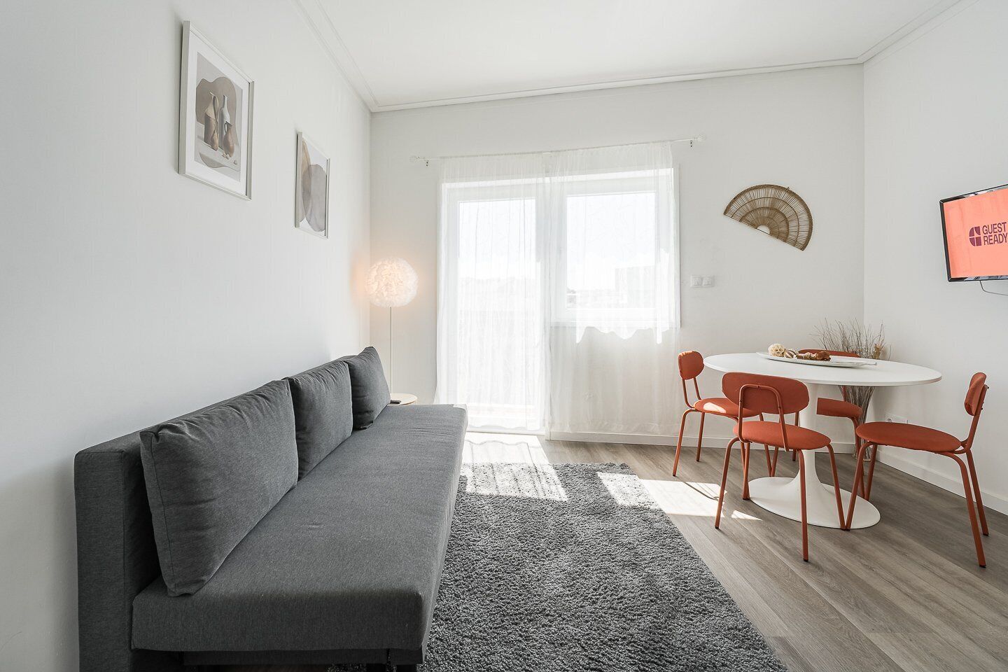 A modern living room with a grey sofa, a round dining table with four chairs, and a large window with sheer curtains.