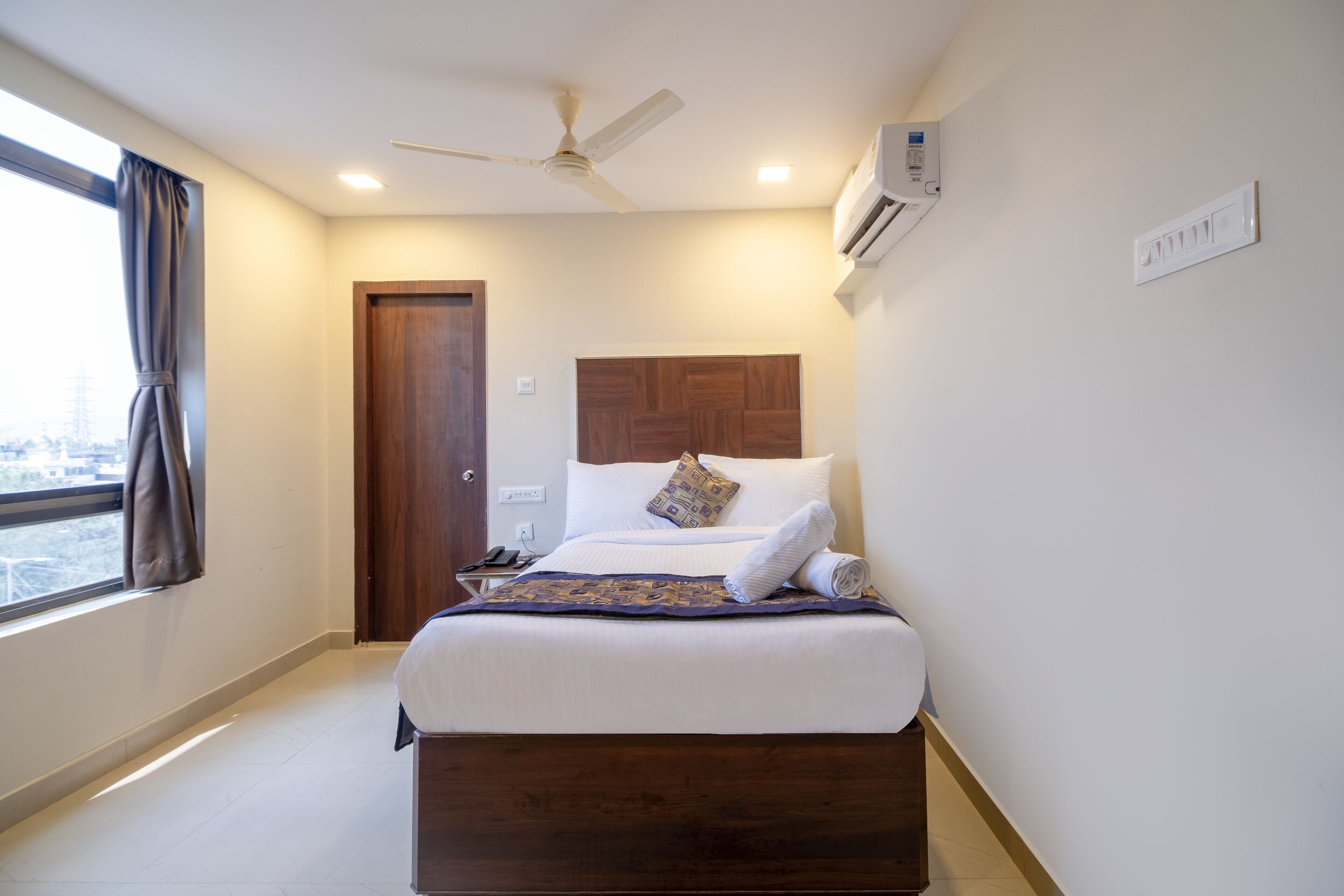 A hotel room with a bed, a ceiling fan, an air conditioning unit, a window with curtains, and a door.