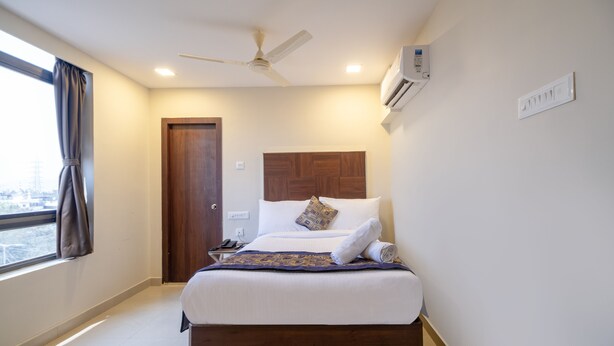A hotel room with a bed, a ceiling fan, an air conditioning unit, a window with curtains, and a door. - Monday Hotels - Swarna's The Capital (Penumuluru)