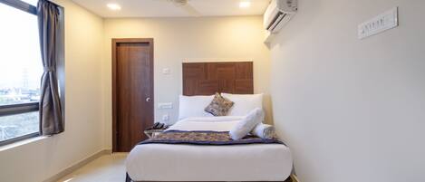 A hotel room with a bed, a ceiling fan, an air conditioning unit, a window with curtains, and a door.