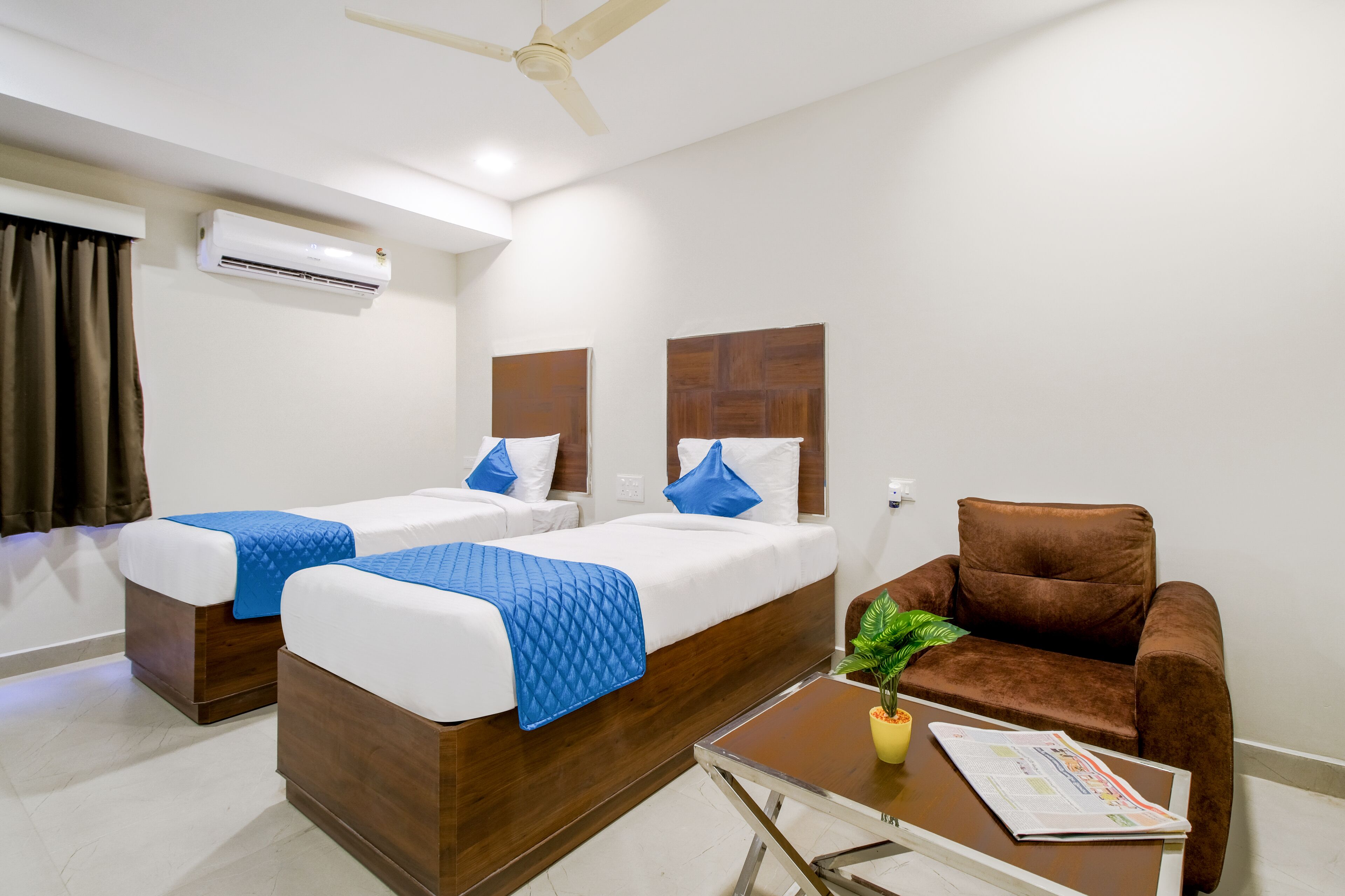 A hotel room with two beds, a brown sofa, a small table with a plant, and an air conditioning unit.