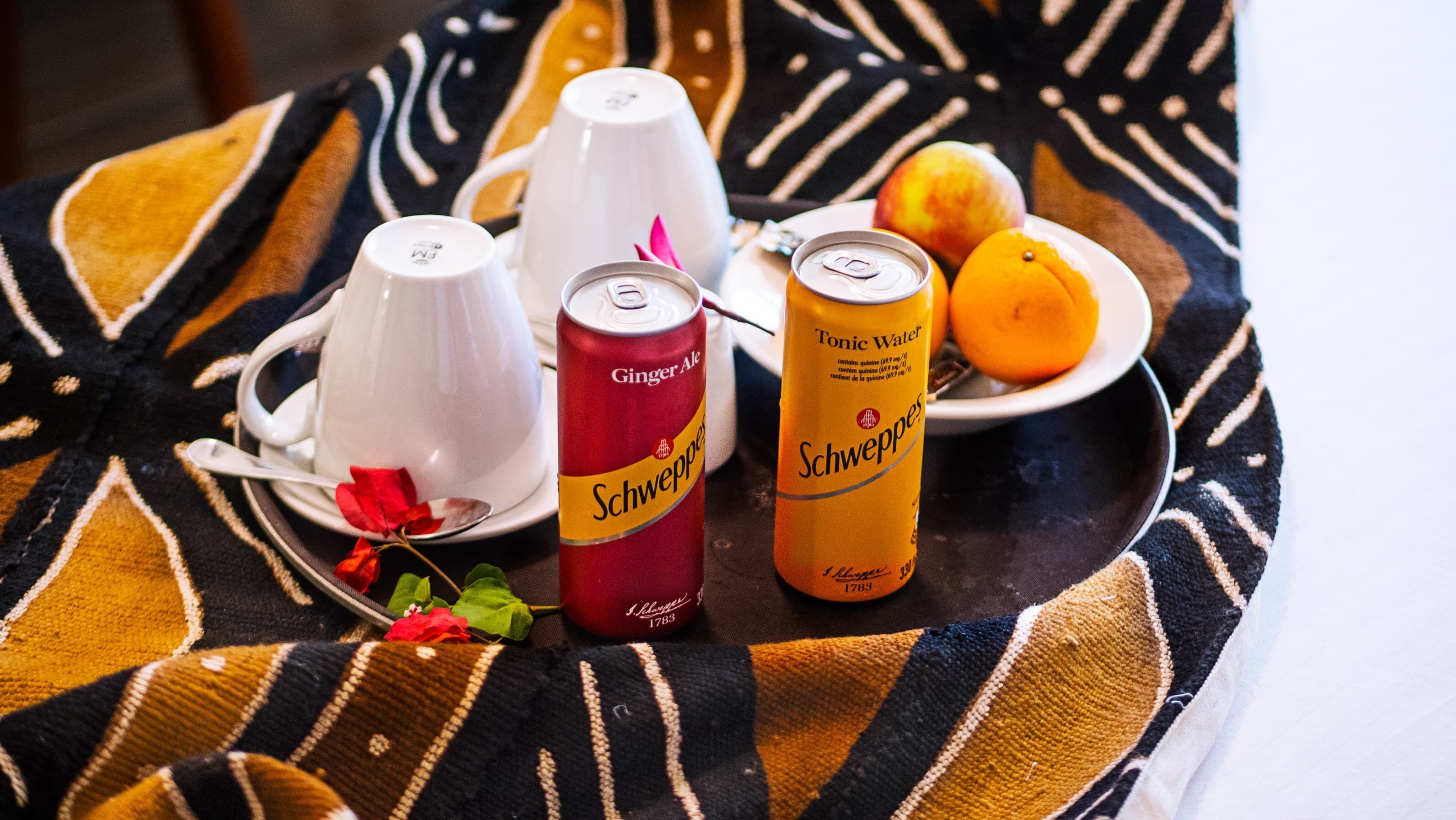 A table with a patterned tablecloth, a white teapot, a red can of ginger ale, a yellow can of tonic water, a white cup, a plate with an orange and a peach, and a spoon with a red flower.