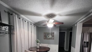 Zona pranzo con ventilatore a soffitto, tavolo con sedie e frigorifero nella cucina adiacente.