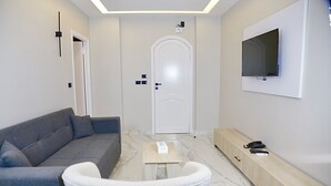 Una sala moderna con un sofá gris, un sillón blanco, una mesa de centro de madera y un televisor montado en la pared.