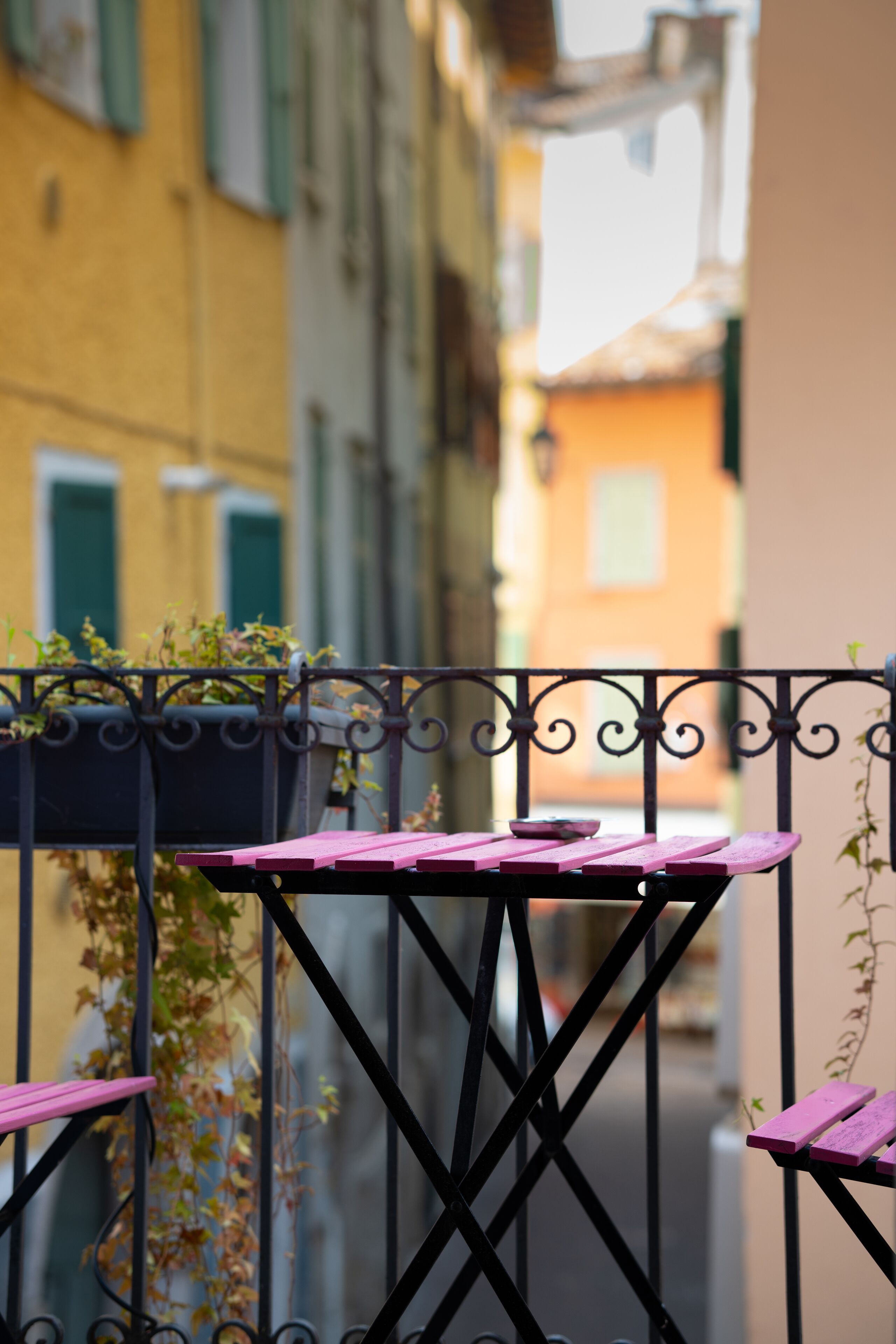 A balcony with a pink table and chairs, surrounded by a wrought iron railing.
