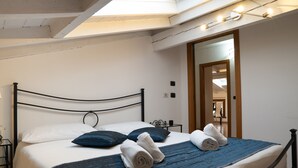 Een net opgemaakt bed met een blauwe sprei en witte lakens, een nachtkastje met een lamp en een dakraam erboven.