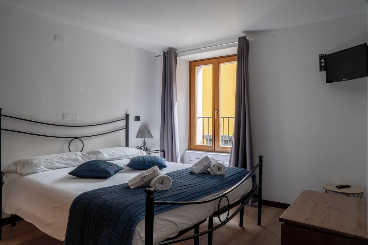 Een keurig opgemaakt bed met een blauwe deken en kussens, een nachtkastje met een lamp, een klein houten tafeltje, en een tv aan de muur.