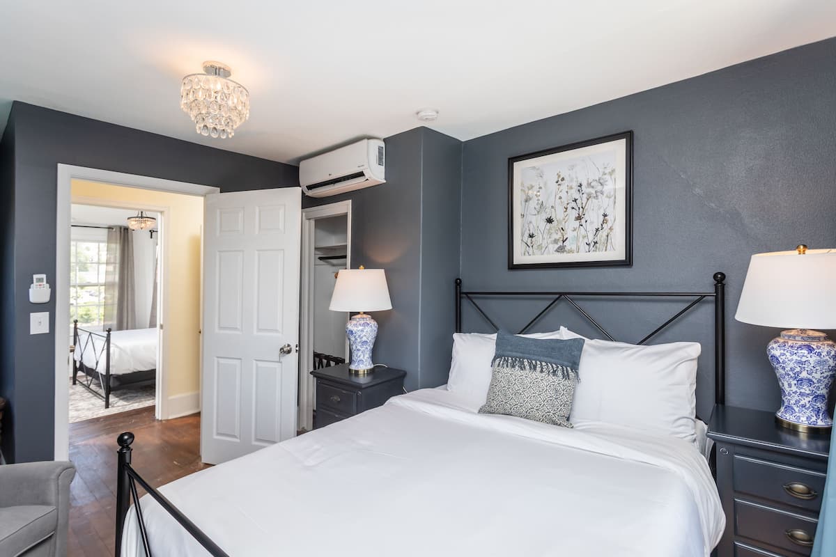 A bedroom with a bed, bedside tables, a chandelier, an air conditioning unit, and a framed artwork on the wall.