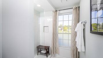 A modern bathroom with a glass shower enclosure, a small wooden stool, a towel rack, and a window with curtains.