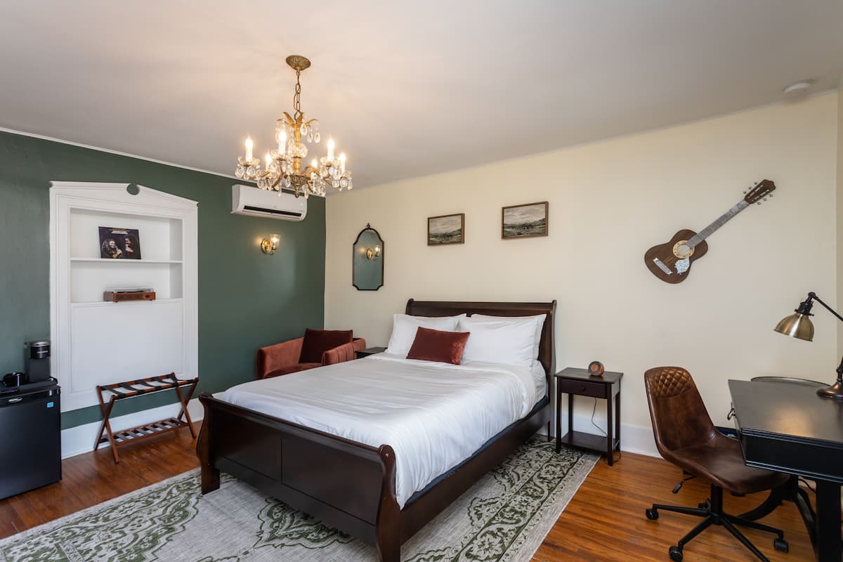 A bedroom with a bed, a desk, a chair, a guitar, a chandelier, and a small shelf with pictures.